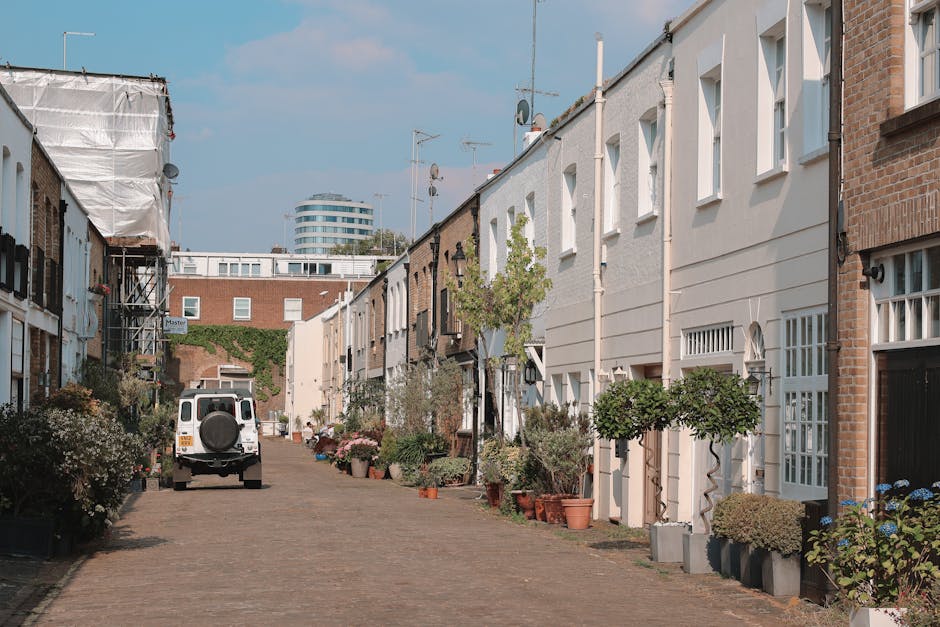A quiet residential street scene in Notting Hill with a white van parked on a cobblestone pavement, as part of a home relocation or furniture transport process. The street is lined with terraced houses featuring white and brick facades, some with front gardens or potted plants near doorways. Several large cardboard boxes and wrapped furniture items are visible on the pavement, ready for loading. A dollop of greenery from small trees and potted plants adds a touch of natural decor. In the background, a larger modern building with rounded architecture contrasts with the traditional houses. The scene is well-lit with daylight, emphasizing the logistics involved in packing and moving services, with the Notting Hill Removals team likely engaged in the loading process at this location during a house removal or a quick emergency move.