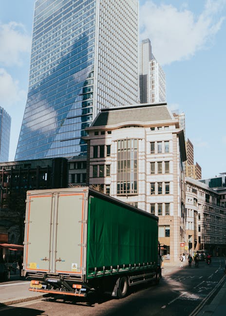 A large green moving truck is parked on a city street in front of tall modern glass skyscrapers and older stone buildings, with the back doors closed and secured with metal latches. The vehicle is positioned near the curb, and there are a few pedestrians walking along the sidewalk nearby. The scene depicts a typical urban environment suitable for house removals and furniture transport during a home relocation, as serviced by Notting Hill Removals. The sky is partly cloudy, providing natural lighting that highlights the reflective surfaces of the surrounding buildings. The truck is ready for loading or unloading, with potential interior space for packed boxes and furniture to facilitate packing and moving processes in the context of emergency same-day removals in Notting Hill.