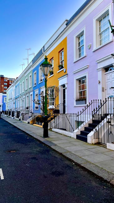 A quiet residential street scene in Notting Hill with a white van parked on a cobblestone pavement, as part of a home relocation or furniture transport process. The street is lined with terraced houses featuring white and brick facades, some with front gardens or potted plants near doorways. Several large cardboard boxes and wrapped furniture items are visible on the pavement, ready for loading. A dollop of greenery from small trees and potted plants adds a touch of natural decor. In the background, a larger modern building with rounded architecture contrasts with the traditional houses. The scene is well-lit with daylight, emphasizing the logistics involved in packing and moving services, with the Notting Hill Removals team likely engaged in the loading process at this location during a house removal or a quick emergency move.