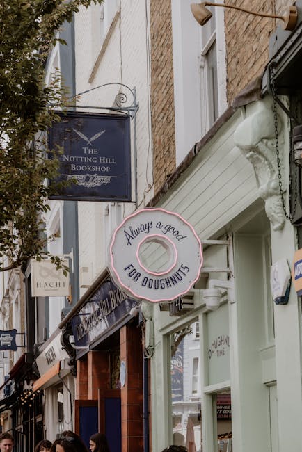 Photograph of a narrow street with a brick building facade hosting multiple shop signs, including a blue sign for 'The Notting Hill Bookshop' and another for a doughnut shop with a large, round, light pink sign featuring a graphic of a doughnut and the message 'Always a good for doughnuts.' In front of the shops, a sidewalk area is visible with people walking, some partially obscured by the foreground objects. The scene captures the exterior environment during daytime, with natural light illuminating the building fronts and signage. The overall setting suggests a retail or commercial area within a residential neighbourhood, typical of the Notting Hill district, with potential for home relocation or moving logistics services offered by companies like Notting Hill Removals being relevant to the context.