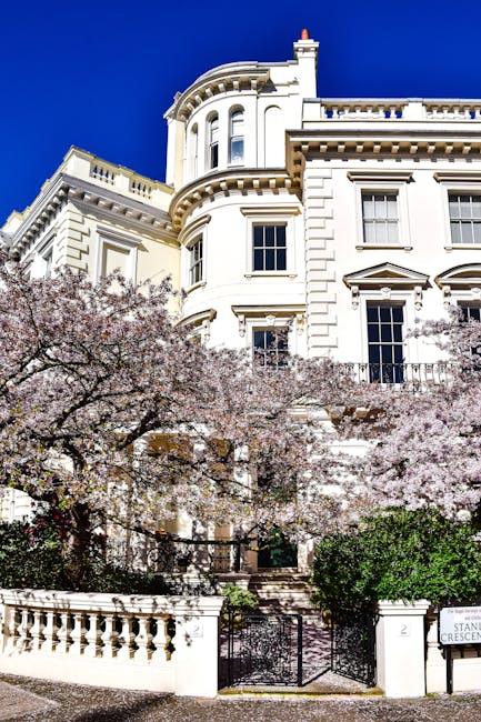 A large, elegant white Victorian-style house with ornate architectural details, multiple windows, and a curved corner tower situated in a residential area. In front of the property, there is a stone retaining wall with a decorative balustrade and a set of steps leading up to the entrance. Pink blossom trees with full, flowering branches partially obscure the lower part of the house, adding colour and charm. The driveway and pavement are visible at the front, with a black gate and a small sign indicating the street name. The bright blue sky provides clear lighting, highlighting the building's intricate mouldings and historic character. The setting suggests a quiet, upscale neighbourhood suitable for house relocations or home removals, as managed by [COMPANY_NAME], with an emphasis on careful handling and transport logistics.