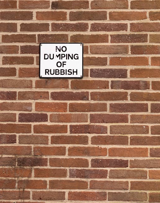 A white rectangular sign mounted on a red brick wall displaying the message 'NO DUMPING OF RUBBISH' in black capital letters, with the text arranged in four lines. The sign is positioned slightly above the center of the image and the bricks feature a traditional horizontal pattern with alternating rows of full and half bricks, creating a textured background. The lighting is natural, casting subtle shadows on the bricks, and the wall appears clean and well-maintained. This scene could be associated with an area where house removals or moving services are offered, reflecting local regulations on waste disposal, as indicated by the page title about Kensington & Chelsea council rules on bulky waste. Notting Hill Removals might help clients comply with such regulations during home relocations involving packing, furniture transport, or clearance of rubbish.