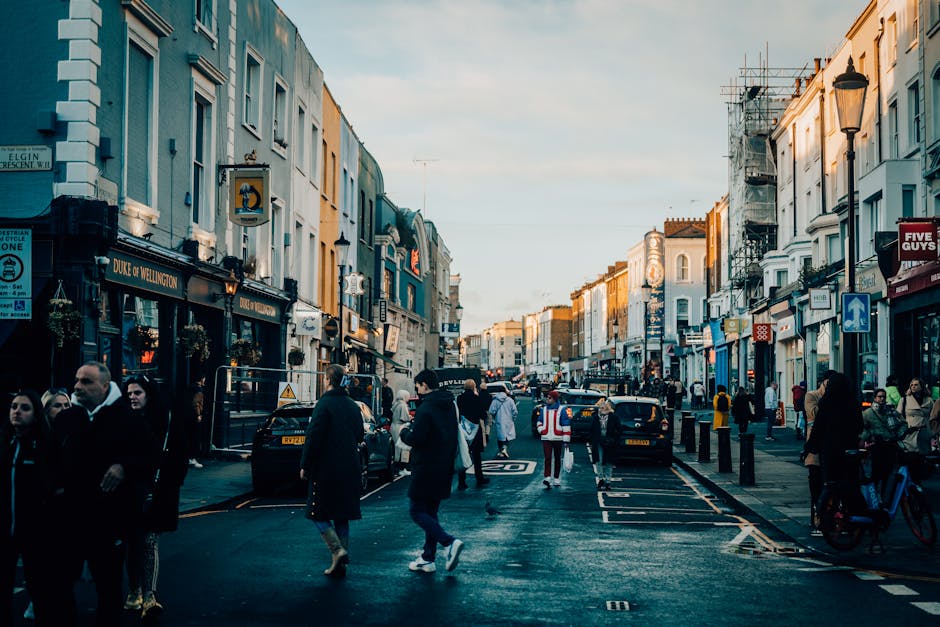 A busy street scene in Notting Hill with pedestrians walking along the pavement, some carrying bags or pushing bicycles, and others waiting to cross the road. The street is lined with multi-storey buildings with various shopfronts, including a pub named Duke of Wellington, and signage visible on the facades. Parked cars are lined along the curb, and a vehicle with a Notting Hill Removals personnel wearing a high-visibility vest is seen near the loading area, indicating a home relocation process. The scene is illuminated by natural daylight, capturing the lively atmosphere typical of a neighborhood where furniture transport and packing are part of ongoing moving or clearance activities related to council rules on bulky waste. [COMPANY_NAME] occasionally appears naturally in the context of loading and transport within this urban environment, reflecting the logistical aspects of removals and home moving services in this residential area.