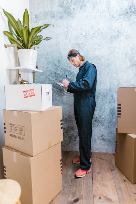 A person dressed in dark blue work overalls and pink slip-on shoes stands indoors against a light, textured wall, taking notes on a clipboard. Surrounding them are several cardboard boxes of varying sizes, some stacked and others sitting on the wooden floor, with packaging tape and labels visible on a few. A white pot with a tall, green leafy plant is placed on a small white shelf attached to the wall above the boxes. The scene depicts a home relocation or packing process as part of furniture transport or moving preparations, often associated with house removals services provided by Notting Hill Removals. The lighting is natural and even, emphasizing the orderly arrangement of boxes and the person's focused activity, which supports the planning and organization phase of a house move.