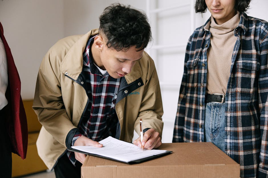 A man in a beige jacket and plaid shirt is leaning over a cardboard box while signing a document on a clipboard, with a pen in hand. A woman, dressed in a beige turtleneck and blue plaid shirt with jeans, stands nearby observing. They are inside a home, with a white ladder-style shelf visible in the background. The scene captures a moment during a home relocation process, with the man likely completing paperwork related to furniture transport and packing services. The cardboard box in front of them is part of the packing materials used for moving household items, and the setting indicates an organized loading area within the property, prepared for the next stage of the move. Notting Hill Removals, specializing in removals, may be involved in this furniture transport and packing process, demonstrating professional handling of home relocation activities.