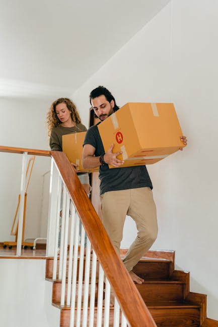 TwoMovers from Notting Hill Removals carrying cardboard packing boxes down a wooden staircase inside a residential property during a home relocation. The movers, a man and a woman, are focused on their task; the man is gripping a large box with both hands while descending the stairs, which are constructed of polished wood with a white-painted wall and a wooden handrail on one side. The woman is positioned slightly behind, holding another cardboard box, ready to assist in the furniture transport process. The interior environment is well-lit with natural light, and the scene is part of a professional packing and moving operation focused on stair and safety solutions in a house moving context, reflecting careful handling of fragile belongings during furniture transport undertaken by [COMPANY_NAME].