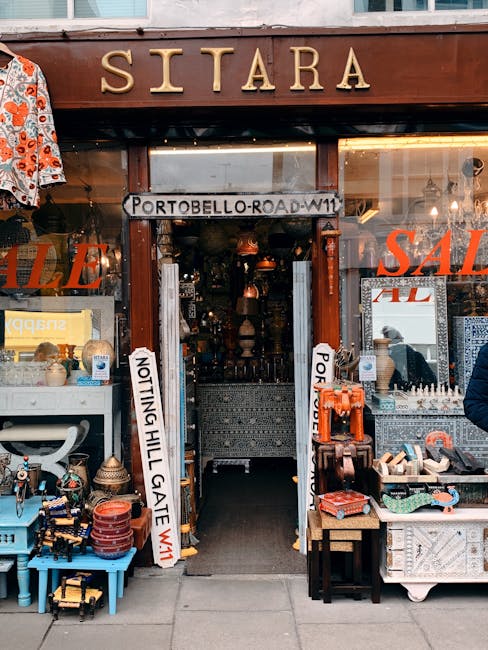 The entrance to a shop named 'SITARA' is visible, with large gold lettering on a wooden sign above the doorway. The shop's storefront includes various items arranged on display tables and shelves, such as ceramic pots, decorative objects, and small furniture pieces. A white sign with black text indicating a street address, 'PORTOBELLO ROAD W11', is placed across the doorway. Two vertical white banners with black text—'NOTTING HILL GATE' and 'PORTOBELLO ROAD'—are positioned on either side of the entrance, suggesting a busy shopping area. Inside the shop, glimpses of pendant lights and hanging ornaments can be seen, with warm lighting illuminating the space. Outside, on the pavement, a small black trolley or cart is available for loading or unloading, indicating arrangements for house removals or furniture transport. This setting is typical for moving antiques or household items as part of a home relocation process, with [COMPANY_NAME] potentially involved in managing the logistics and safe packing for the move.