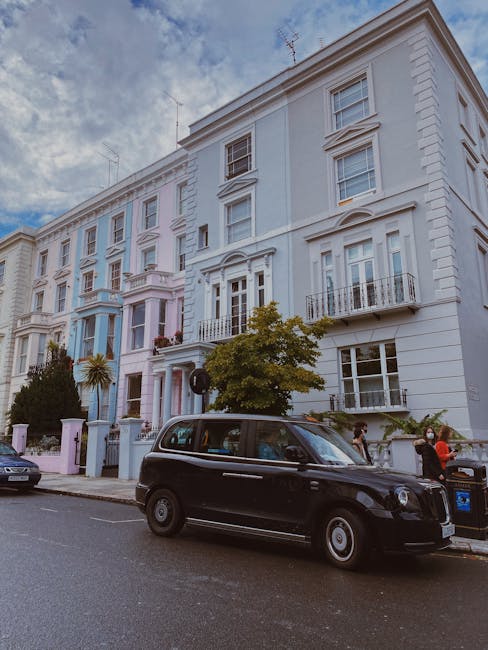 A black London taxi is parked on the street in front of a row of residential Victorian-style terraced houses with ornate white facades, large windows, and small balconies. The houses are painted in pastel shades, with some featuring pink and blue accents. A tree with green foliage partially obscures one of the buildings. Two people, one wearing a face mask, are standing on the pavement near the taxi, possibly preparing for a home relocation or furniture transport. The scene is set during daylight with partly cloudy skies overhead. This image captures the typical street environment for house removals and moving services in Notting Hill, with elements such as loading equipment or boxes not visible but implied by the context of relocation provided by Notting Hill Removals.
