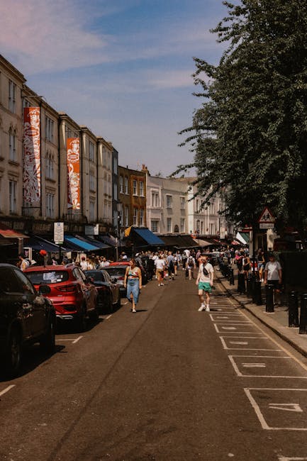 The image depicts a busy street scene in Notting Hill during daylight hours, with a row of multi-storey buildings lining the pavement on the left side. Several parked cars are visible along the curb, with some partially obscured by pedestrians walking in both directions. On the right side, there is a designated loading area marked by white painted lines on the asphalt, with black bollards separating it from the pedestrian walkway. Large trees with dense foliage overhang the scene, casting shadows on the pavement. In the background, people are engaged in various activities, and the sky is partly cloudy with patches of blue. This setting could illustrate the typical environment where [COMPANY_NAME], such as Notting Hill Removals, may assist with home relocation or furniture transport, particularly when navigating streets requiring parking permits for moving day.
