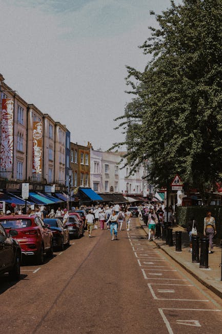 A busy street scene showing a row of multi-storey buildings with commercial storefronts at ground level, some with blue awnings, and others with signs and window displays. Parked cars line the left side of the street, including a red vehicle in the foreground. Pedestrians are walking along the pavement and across the street, with some carrying boxes and bags, possibly indicating moving activities. A large tree with lush green foliage extends over the right side of the street, partially shading the sidewalk. The sky is clear and bright, suggesting daytime. Inside a property, a home relocation or furniture transport process is underway, with packing materials such as cardboard boxes and plastic wrap visible near the building entrance. A moving trolley, straps, and blankets may be present to support the loading process. This scene reflects the logistical aspects of house removals and the variety of packing and transport preparations involved, aligning with the information discussed in the page titled 'Why Notting Hill removals quotes vary and how to compare' from Notting Hill Removals.