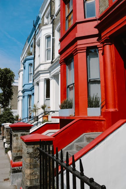 A row of three Victorian-style terraced houses on a residential street, featuring white, blue, and red facades with large bay windows and decorative architectural details. The white house has a black wrought iron fence and stone steps leading up to the front door, with potted plants on the staircase. The blue house is set slightly back, with similar stone steps and a small garden area. The red house has a prominent front entrance with a wide red staircase, two rectangular planters with greenery on either side of the doorway, and a black metal railing along the staircase. The street is illuminated by bright daylight with a clear blue sky, and there are some trees partially visible in the background. This scene may be part of a property examination for home relocation or furniture transport, with a focus on building facades and entryways indicative of residential removals services on a typical urban street, as provided by Notting Hill Removals.